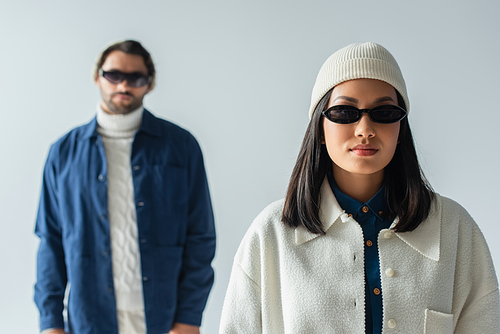 young asian woman in dark sunglasses and white jacket looking at camera near blurred man isolated on grey