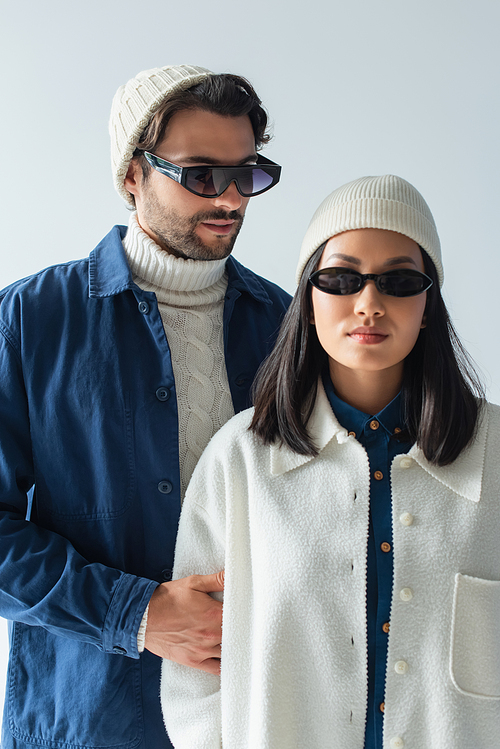 young asian woman in white beanie and dark sunglasses near man in blue jacket isolated on grey