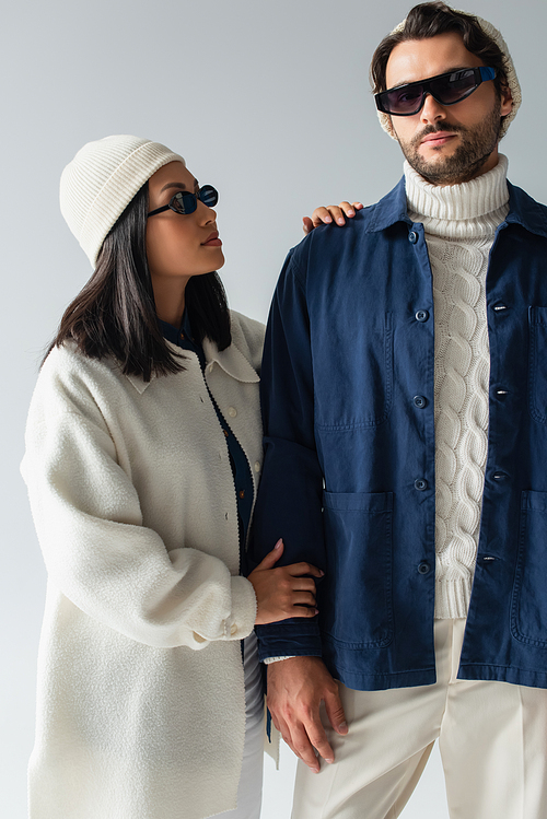 asian woman in white jacket and dark sunglasses looking at stylish man isolated on grey