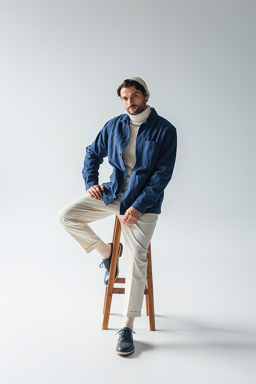 full length view of man in blue jacket and white trousers sitting on high stool on grey