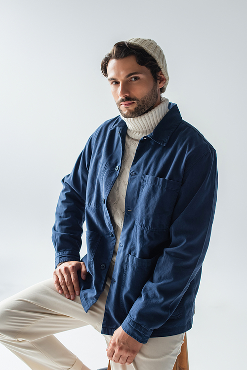 stylish man in blue jacket and white beanie looking at camera isolated on grey