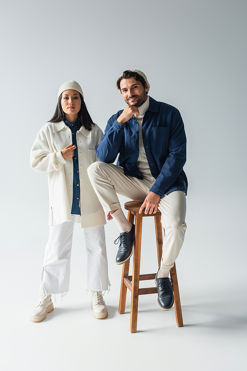 full length view of trendy asian woman near smiling man sitting on high stool on grey