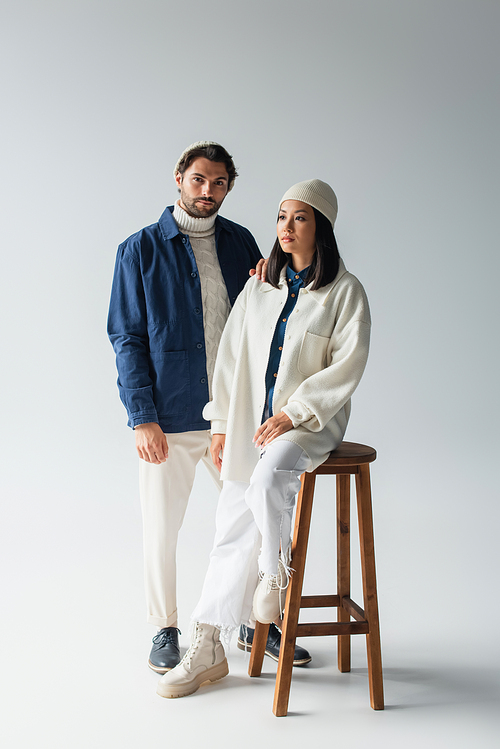 stylish man looking at camera near asian woman sitting on high stool on grey