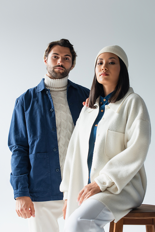 trendy man looking at camera near asian woman in white jacket and beanie isolated on grey