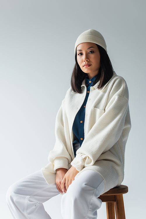 pretty asian woman in white beanie and jacket sitting on stool and looking at camera isolated on grey