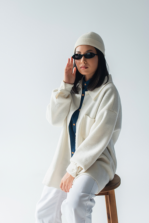 young asian woman in white jacket and beanie touching dark sunglasses isolated on grey