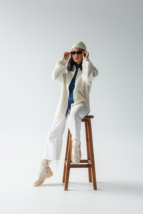 full length view of asian woman in white clothes and black sunglasses posing on high stool on grey