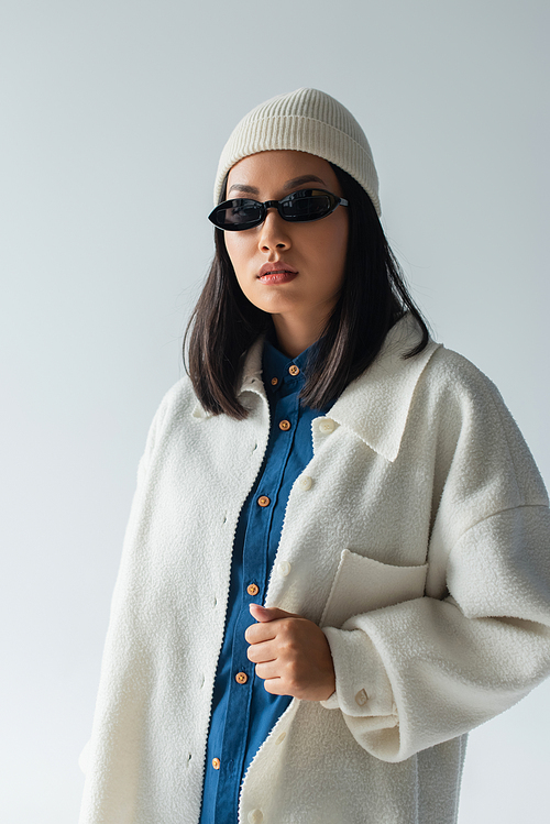 young asian woman in white beanie and black sunglasses looking at camera isolated on grey