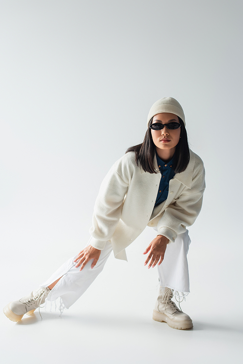 full length view of asian woman posing in white clothes and dark sunglasses on grey