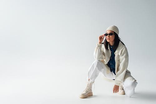 full length view of asian woman in white clothing adjusting sunglasses and looking away on grey