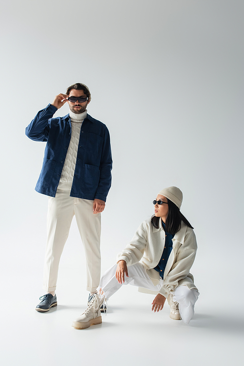 full length view of trendy man adjusting dark sunglasses near asian woman on grey