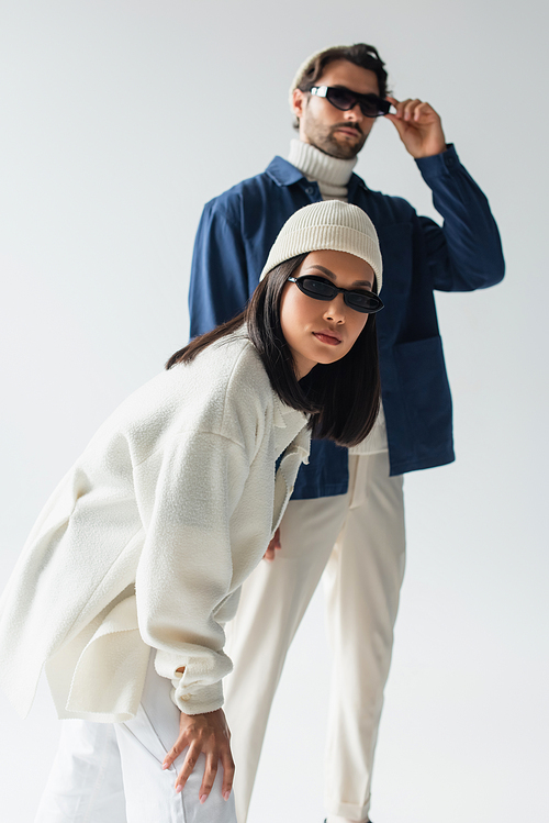 asian woman in white jacket and dark sunglasses looking at camera near blurred man on grey