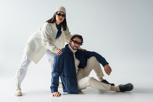 fashionable interracial couple in dark sunglasses looking at camera while posing on grey