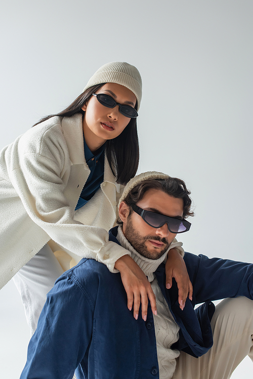 fashionable interracial couple in dark sunglasses and beanies looking at camera isolated on grey