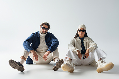 full length view of multiethnic couple in trendy clothes and dark sunglasses sitting on grey