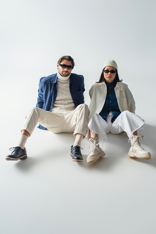 full length view of fashionable interracial couple in dark sunglasses sitting on grey