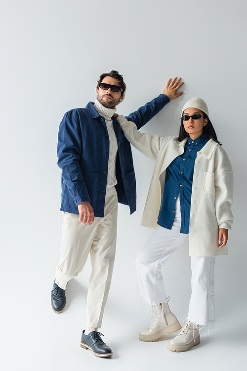 full length view of stylish interracial couple in white and blue clothes and sunglasses on grey