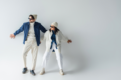 full length view of interracial couple in trendy clothes and sunglasses posing on grey