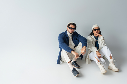 full length view of stylish interracial couple in dark sunglasses sitting on grey with copy space