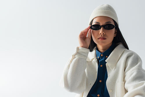 young asian woman in white jacket and beanie adjusting dark sunglasses isolated on grey