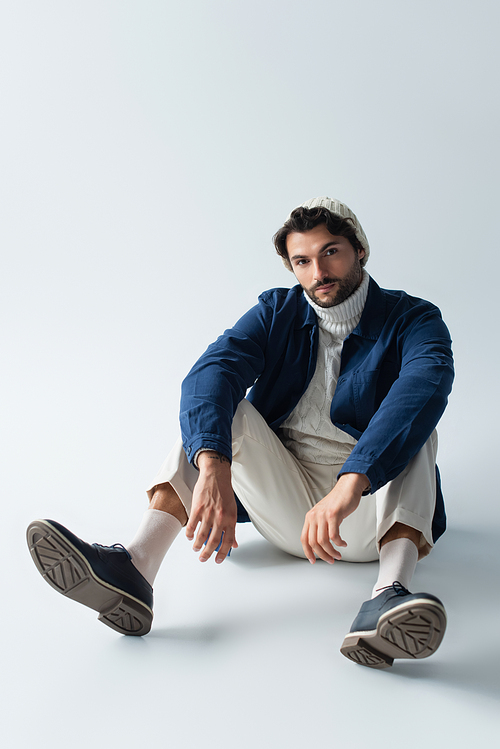 full length view of brunette man in fashionable clothes sitting on grey