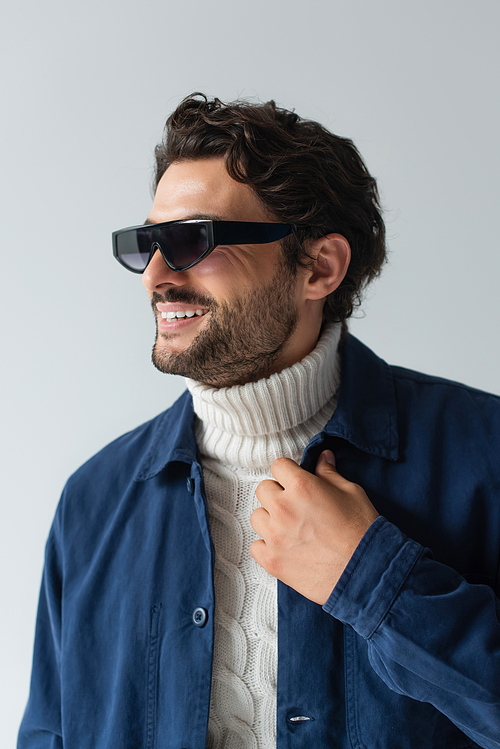 happy man in blue jacket, white sweater and dark sunglasses looking away isolated on grey