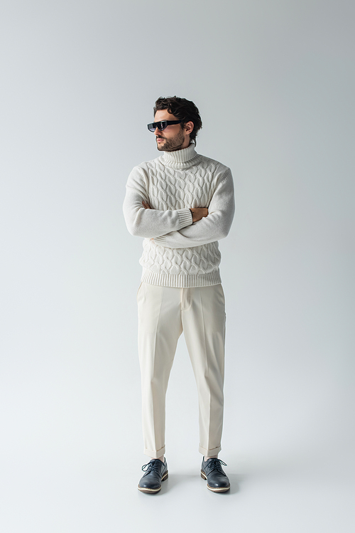 brunette man in white trousers, knitted sweater and dark sunglasses standing with crossed arms on grey