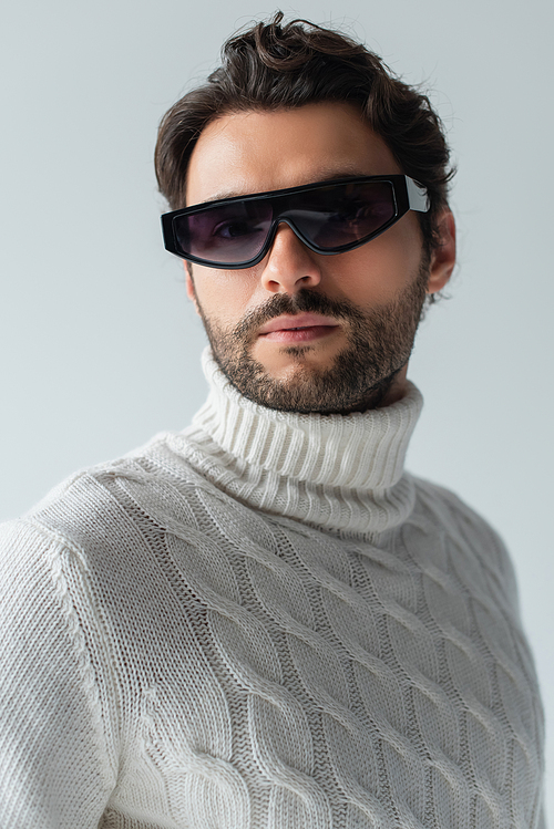 trendy man in white sweater and dark sunglasses looking at camera isolated on grey