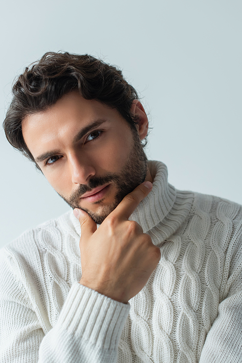 brunette man in white knitted sweater touching chin and looking at camera isolated on grey