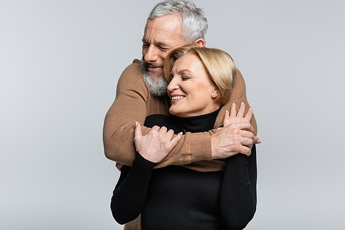 Mature man with closed eyes hugging cheerful wife isolated on grey