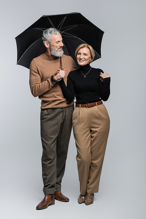 Positive mature woman looking at camera near stylish husband with umbrella on grey background