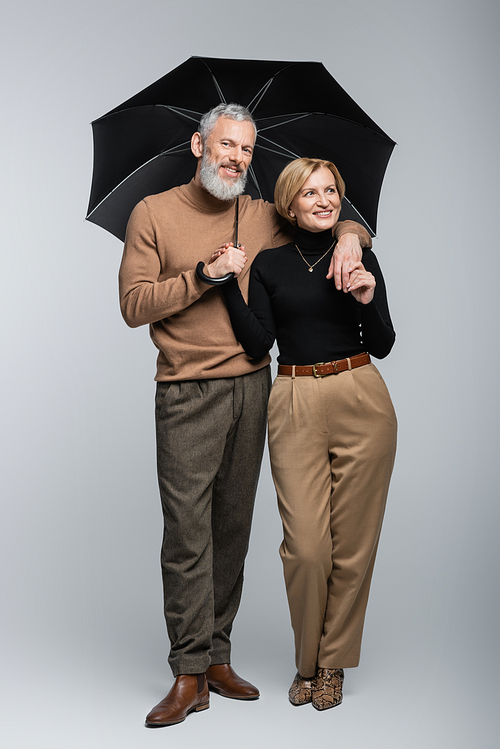 Stylish mature man holding umbrella and hugging cheerful wife on grey background