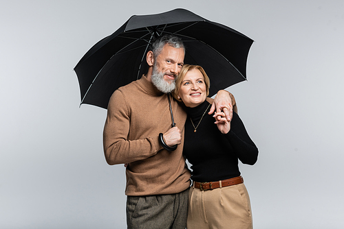 Positive and stylish mature couple holding umbrella and hands isolated on grey