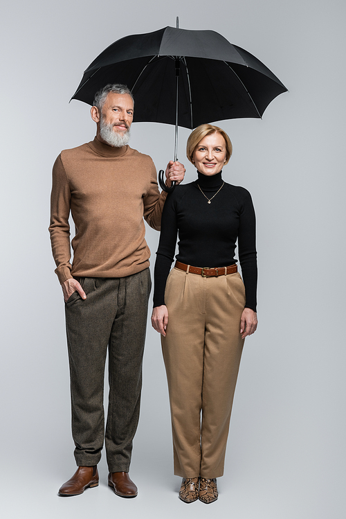 Full length of stylish mature couple with umbrella looking at camera on grey background