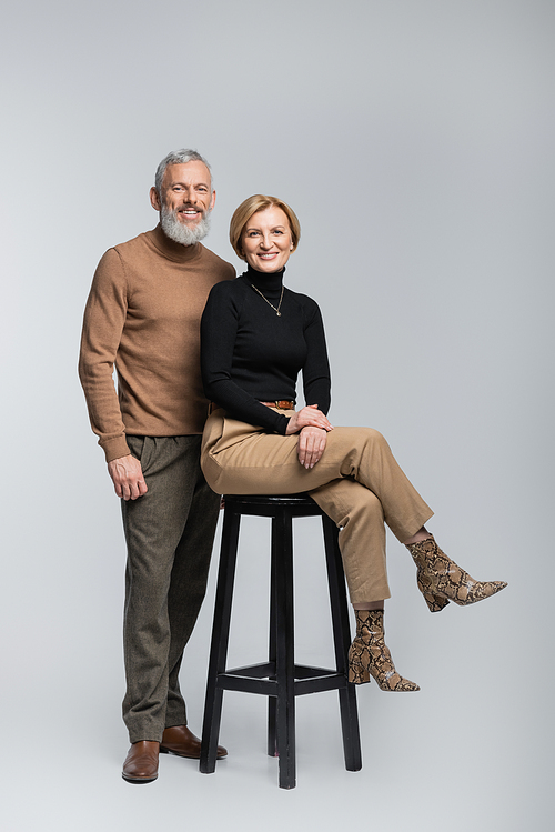 Smiling stylish man looking at camera near wife sitting on chair on grey background