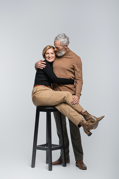 Cheerful stylish woman hugging mature husband while sitting on chair on grey background