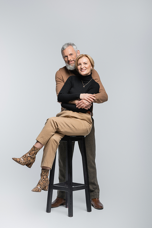 Positive mature man embracing trendy wife sitting on chair on grey background