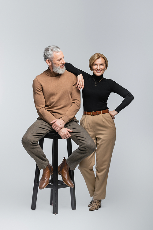 Smiling mature woman posing near stylish husband sitting on chair on grey background