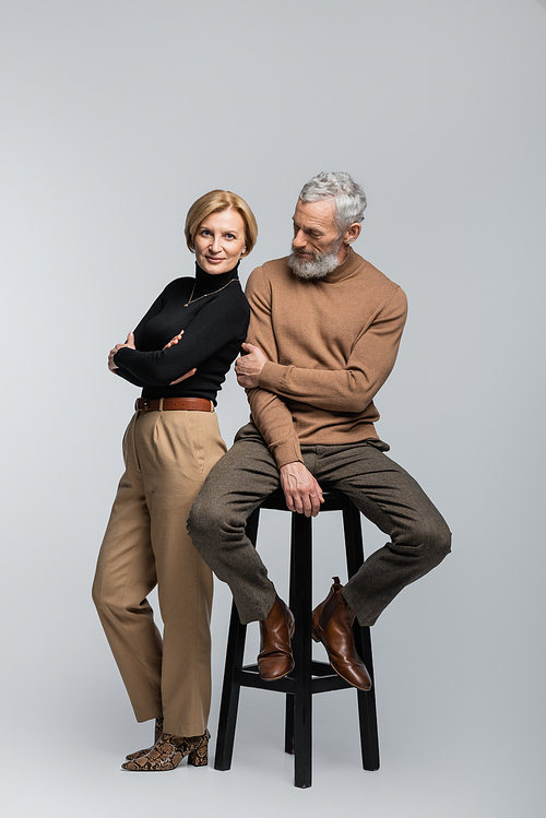 Full length of stylish woman crossing arms near mature husband sitting on chair on grey background