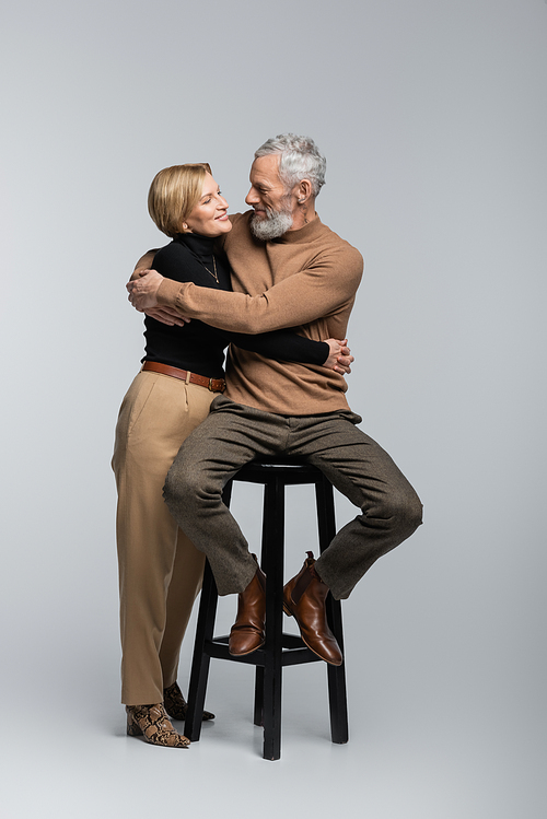 Mature woman hugging stylish husband sitting on chair on grey background