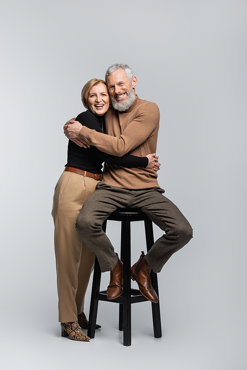 Happy and trendy mature couple hugging and looking at camera on grey background