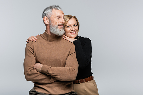 Smiling mature woman in turtleneck hugging husband and looking at camera isolated on grey