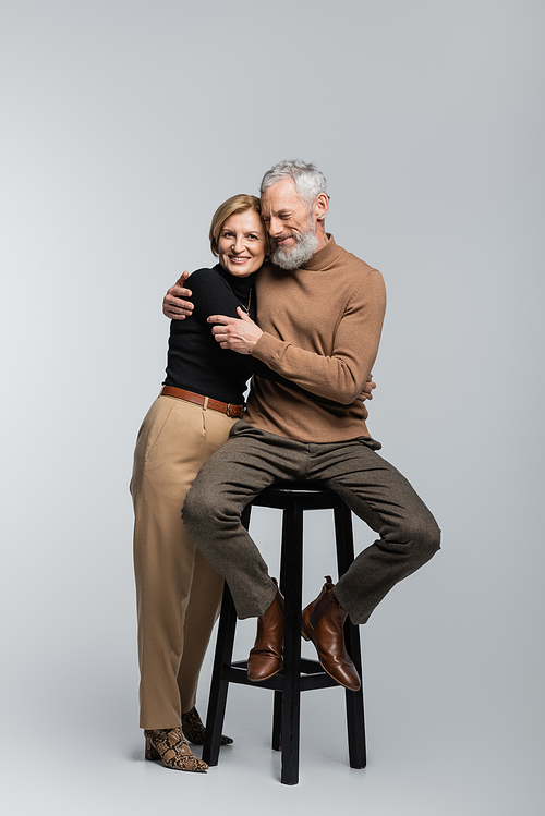 Smiling mature woman hugging husband sitting on chair on grey background