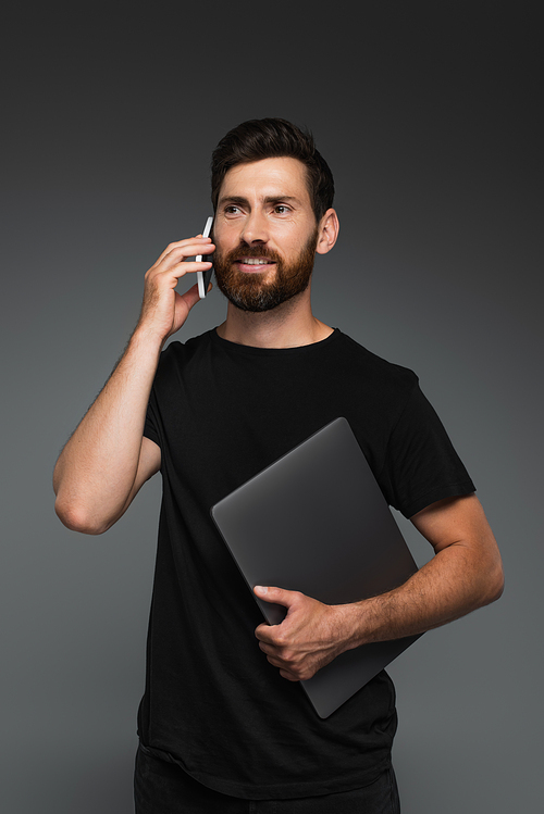happy bearded man in black t-shirt holding laptop while talking on smartphone isolated on grey