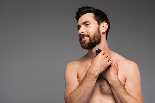 cheerful and shirtless man with hair on chest shaving with electric razor isolated on grey