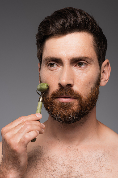 portrait of shirtless man with beard using jade roller while massaging face isolated on grey