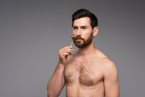 shirtless man with beard using jade roller while massaging cheek isolated on grey