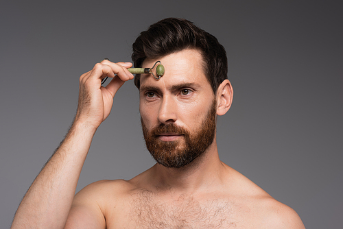 shirtless man with beard using jade roller while massaging forehead isolated on grey