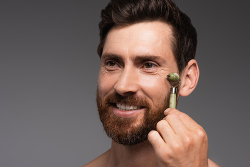 happy man with beard using jade roller while massaging face isolated on grey
