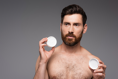 bearded and shirtless man holding container with face cream isolated on grey